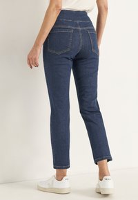 Mörkblå denimbyxor med hög midja och rak benlängd, med bakfickor och kontrasterande gula sömmar. Matchas med vita sneakers.