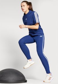 Plava sportska majica s tri bijele pruge, odgovarajuće plave tajice i bijele tenisice s pastelni naglascima, prikazano u aktivnom stavu.