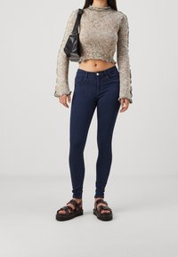 Top de ganchillo en beige y azul, jeans skinny de tiro alto en azul oscuro, y sandalias negras con tiras y suela gruesa.