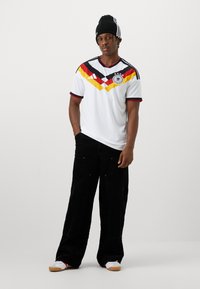 Jeune homme portant un maillot de football d'Allemagne, un bonnet noir, un pantalon noir ample et des baskets blanches, debout devant un fond uni.