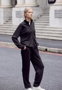 Schwarzes Zip-Jackett und Hosen-Set aus glattem, dehnbarem Stoff. Mit seitlichen Taschen und entspannter Passform, kombiniert mit weißen Sneakern.