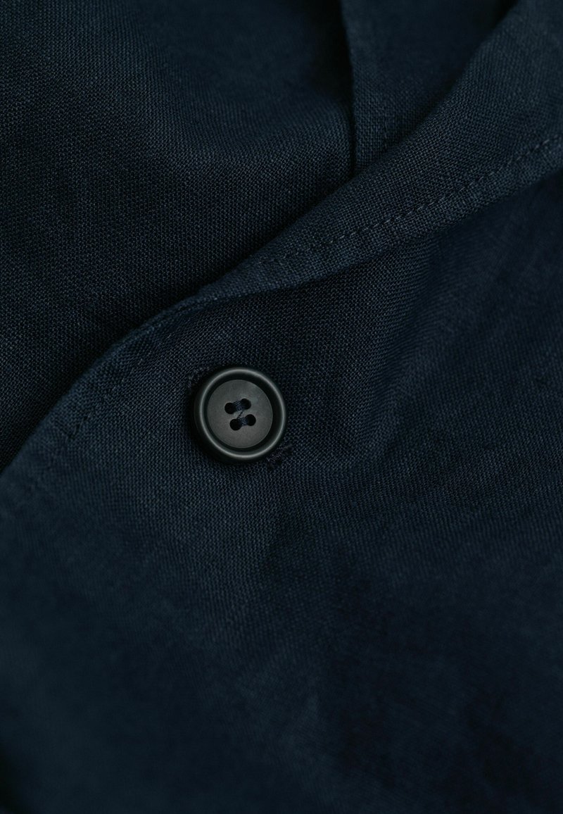 Close-up van een donkerblauwe stof met een genaaide zwarte knoop en zichtbare stiksels langs de gevouwen rand van het kledingstuk.