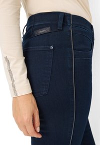 Jean bleu foncé avec une coupe ajustée, des détails de couture subtils et une poche arrière. Fabriqué en denim avec une texture lisse.