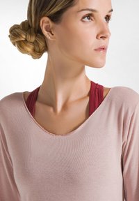 Helles rosa Langarmshirt mit weit ausgeschnittenem Hals, über einem kastanienbraunen Sport-BH getragen; glatte Textur und lockere Passform. Subtile Nähdetails.