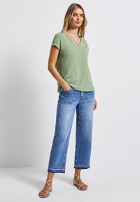 Top verde chiaro a maniche corte con scollo a V, abbinato a jeans in denim blu a gamba larga. Il tessuto sembra avere una consistenza texturizzata, con una vestibilità casual.