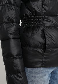 Svart puffjacka med en blank yta, med insvängd midja och ribbade ärmar för en figursydd passform. Lätt och isolerad.