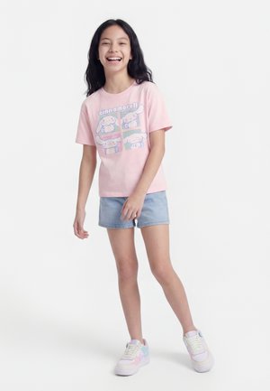 Pige smilende og stående, iført en lyserød t-shirt med Cinnamoroll-grafik, lyseblå denimshorts og sneakers i pastelfarver.