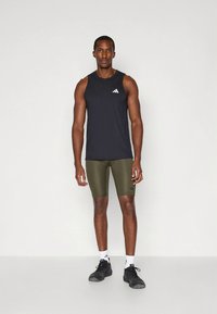 Musta hihaton urheilupaita, jossa on pieni valkoinen logo, yhdistettynä oliivinvihreisiin istuviin shortseihin, mustiin urheilukenkiin ja valkoisiin sukkiin.