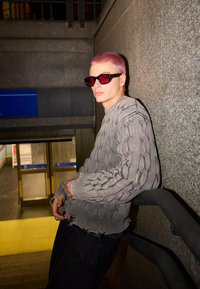 Siv pulover s pletenim vzorcem in raztrganimi robovi, temne kavbojke in črne sončne očala z rdečimi lečami, naslonjen ob strukturirano steno v podzemni železnici.