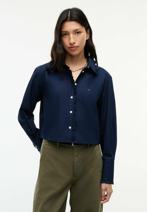 Vrouw met lang donker haar draagt een marineblauw overhemd met knopen en klein logo en olijfgroene broek, staand tegen een effen lichte achtergrond.
