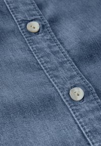 Camicia in denim di colore azzurro chiaro con una texture liscia, caratterizzata da due bottoni avorio e cuciture a contrasto lungo la patta.