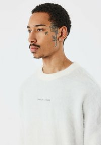 Witte trui met een zachte textuur, ronde hals en geborduurd logo. Beschikt over een slim fit en ribbelkraag, wat een minimalistisch design benadrukt.