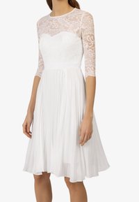 Robe blanche avec corsage en dentelle et manches trois-quarts. La jupe présente des plis, créant une silhouette fluide. Tissu léger et texturé.