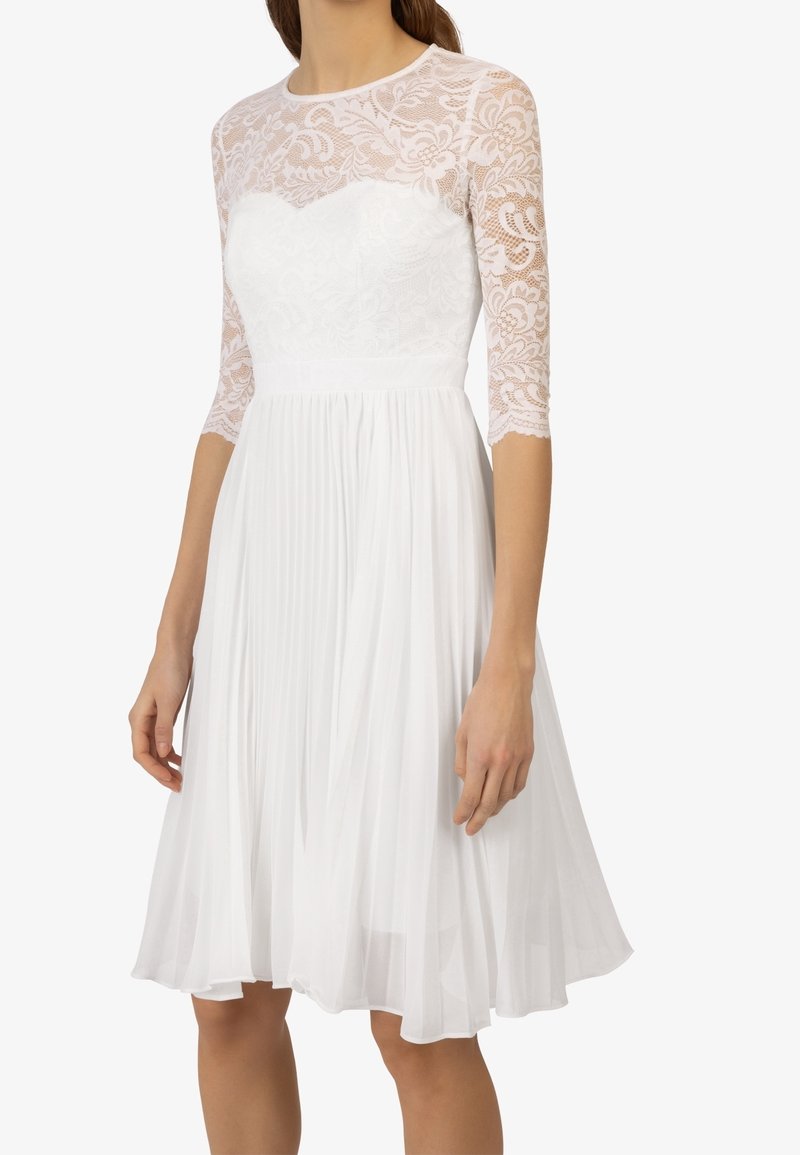 Robe blanche avec corsage en dentelle et manches trois-quarts. La jupe présente des plis, créant une silhouette fluide. Tissu léger et texturé.