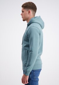 Gai teal krāsas kapucīna džemperis ar priekšējo kabatu, ribotu aproču un apakšmalu; mīksta tekstūra, pieguļošs dizains, valkāts ar ziliem džinsiem.