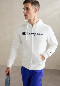 Hvit zip-opp hettegenser med snorhette, med en stor marineblå "Champion"-logo på brystet og en liten logo på ermet.