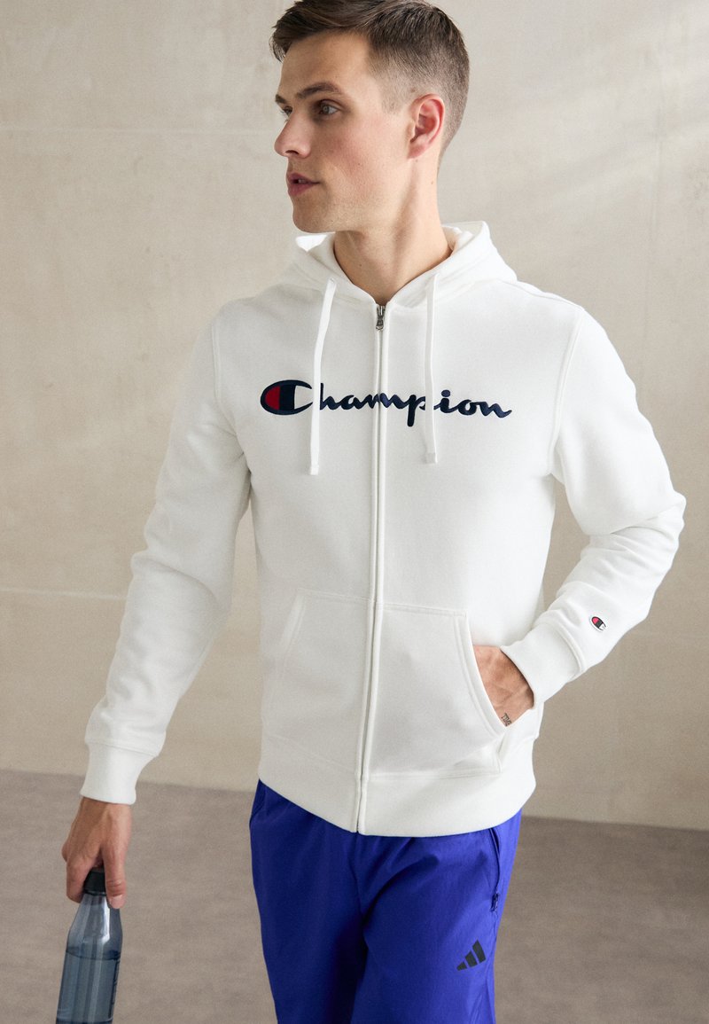 Hvit zip-opp hettegenser med snorhette, med en stor marineblå "Champion"-logo på brystet og en liten logo på ermet.