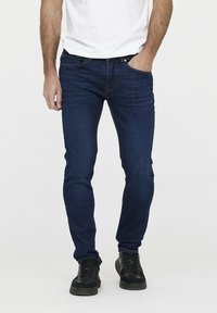 Jeans en denim bleu foncé avec une coupe slim, dotés de poches avant, de passants de ceinture et de détails de couture subtils, associés à des baskets noires.