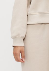 Hånd som hviler ved siden av, iført en beige langermet topp og matchende beige bukser med ribbede mansjetter og midjebånd.