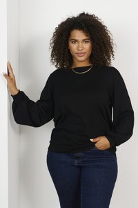 Sweater noir avec des manches bouffantes côtelées et un col rond. Associé à un jean bleu foncé, présenté sur un fond neutre.