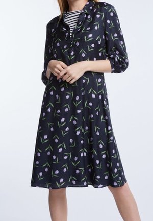 Kvinde iført en knælængde sort kjole med lilla blomsterprint og trekvartærmer, lagdelt over en sort og hvid stribet top.