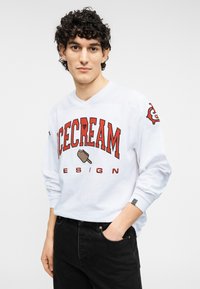 Λευκό πουκάμισο με μακριά μανίκια, με τολμηρό κόκκινο εκτύπωμα "ICECREAM DESIGN" και γραφικό ενός παγωτού σε κώνο. Μαύρο τζιν ολοκληρώνει το ντύσιμο.