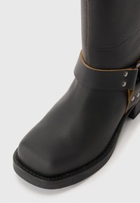 Botín de cuero negro hasta el tobillo con punta redonda, detalle de correa marrón, acentos de hardware plateado y suela de goma texturizada.