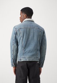 Veste en denim bleu clair avec un col en shearling. Caractéristiques d'une coupe classique, de coutures visibles et de poches latérales, avec un tissu texturé.