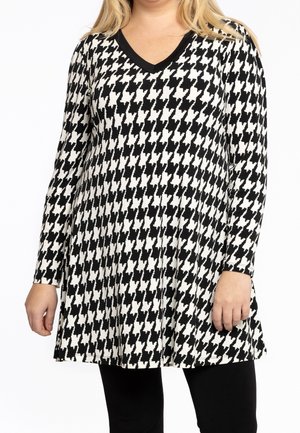 Kobieta ubrana w długą tunikę z czarno-białym wzorem pepitki, z dekoltem w serek, w połączeniu z czarnymi spodniami.