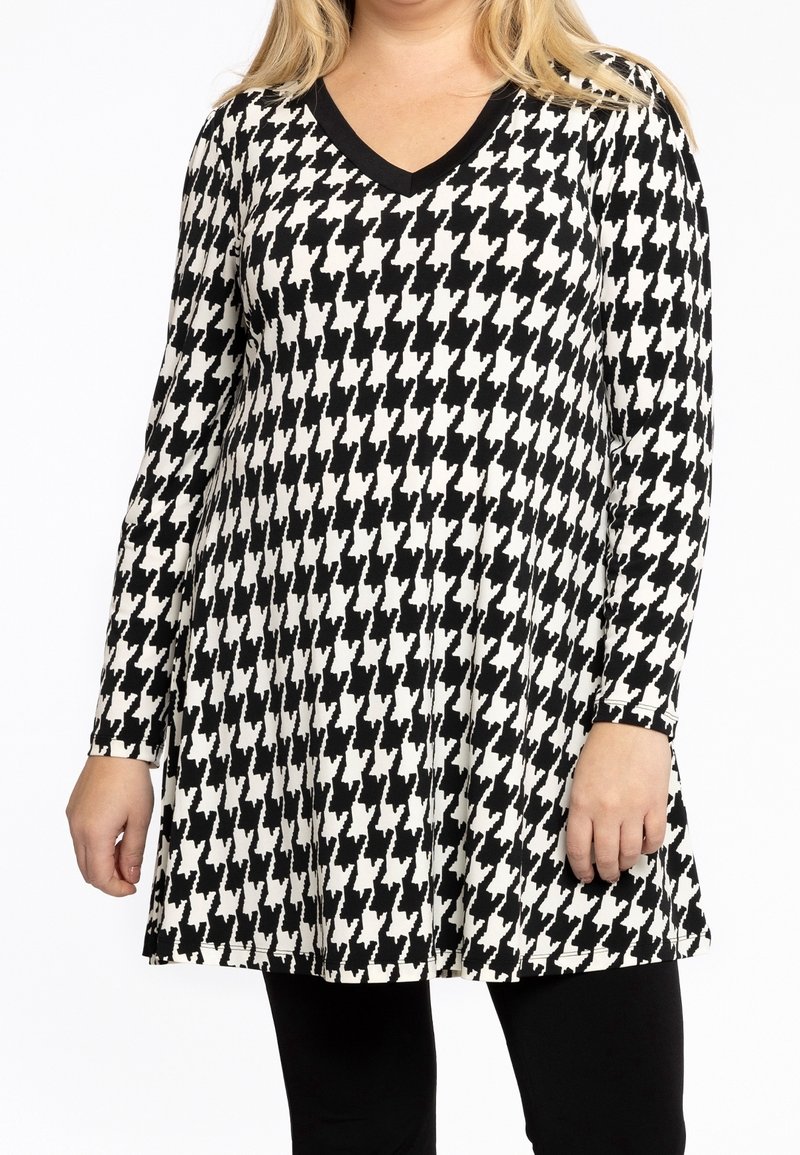 Femme portant une tunique à manches longues à motif pied-de-poule noir et blanc avec un col en V, associée à un pantalon noir.
