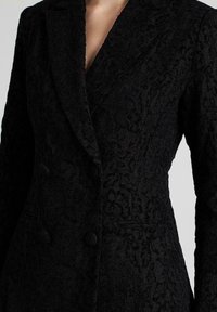 Zwarte dubbelzijdige blazer met een getextureerd bloemenpatroon, gelakte revers en twee voorzakken. Voorzien van grote zwarte knopen.