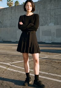 Woman standing with arms crossed in black long-sleeve top, black skirt, black socks, and black sneakers on cracked asphalt near concrete wall.