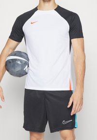 Camiseta deportiva blanca con hombros negros y acentos naranjas, combinada con pantalones cortos negros; incluye logotipos de Nike y un diseño texturizado.