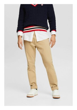 Chinos beige de corte slim, material de algodón, con los puños enrollados, combinados con una camisa blanca de botones y un suéter de punto de cable azul marino con acentos rojos.