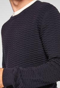 Granatowy sweter o fakturze z wypukłym poziomym wzorem, okrągłym dekoltem oraz kontrastowym kremowym wykończeniem. Ściągacze na mankietach poprawiają dopasowanie i dodają szczegół�ów stylistycznych.