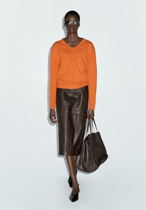Maglione arancione a scollo a V, gonna midi in pelle marrone, scarpe con tacco nere e una borsa a tracolla scura. Texture lisce e design aderente.