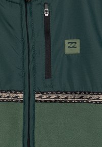 Ciemnozielona kurtka z czarną kieszenią na zamek, wzorzystym wykończeniem i wypukłą naszywką z logo na piersi.