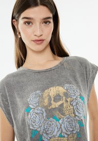 Camiseta gris de corte oversize con una textura deslavada, que presenta un gráfico central de calavera dorada rodeado de rosas azul claro. Cuello redondo, mangas cortas.
