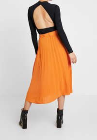 Top nero a maniche lunghe con schiena scoperta abbinato a una gonna midi plissettata arancione. L'outfit è completato da stivaletti neri.