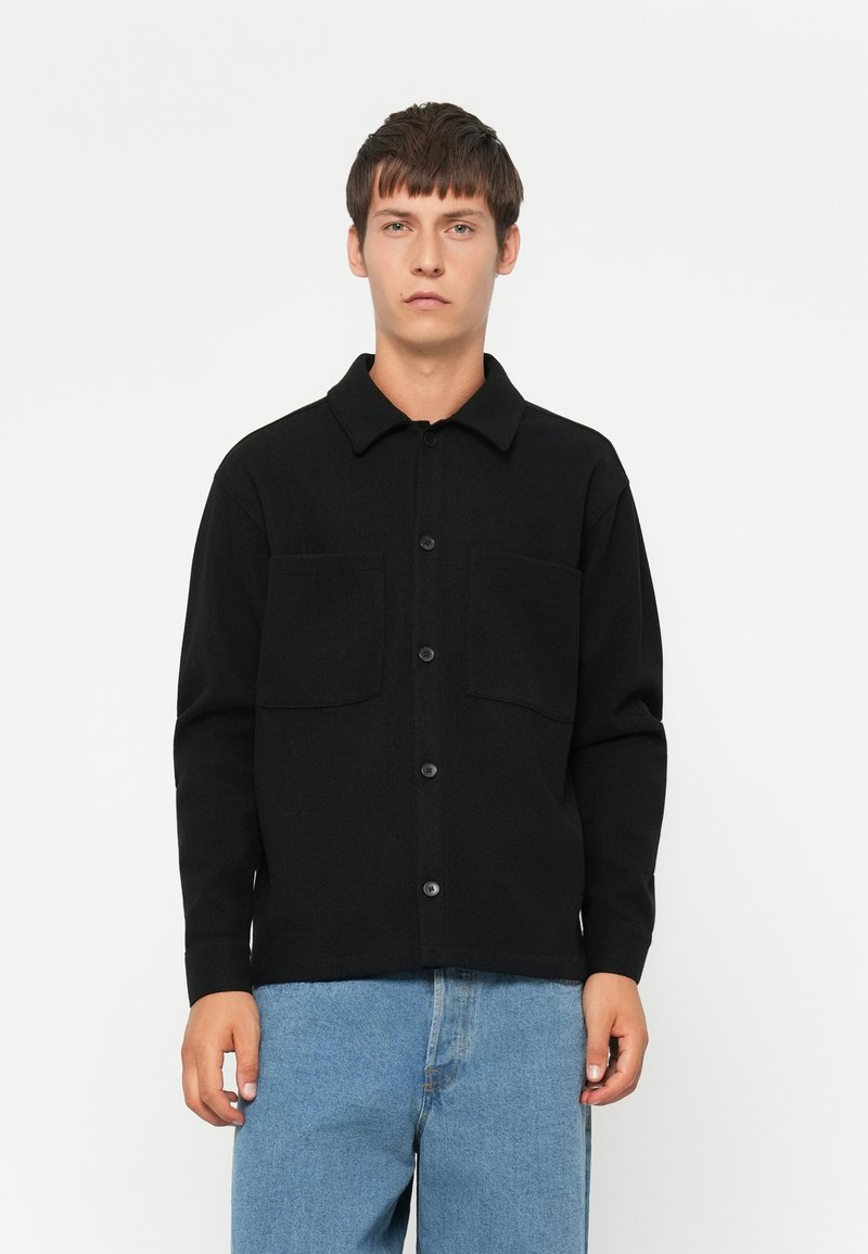 Young man wearing a black buttoned shirt jacket with two chest pockets and light blue jeans against a plain white background.
