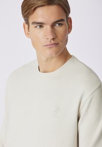 Felpa beige chiaro in morbido misto cotone con scollo rotondo. Presenta un logo USPA in rilievo sul davanti. Texture liscia, vestibilità comoda.