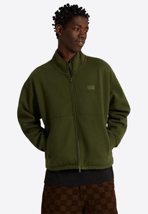 Jonge man met korte twists, draagt een olijfgroene jas met rits en bruine geruite broek, handen in de zakken, kijkt zijwaarts en lacht.