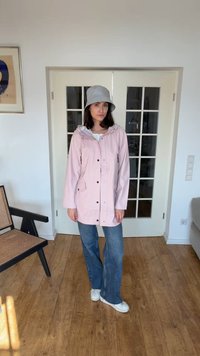 Kvinne iført en lys rosa regnjakke, blå jeans, hvite joggesko og en grå bøttehatt som står innendørs på tregulvet.