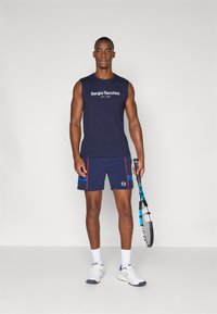 Marineblå ærmeløs skjorte med hvidt logo, parret med marineblå shorts med røde detaljer. Holder en tennisracket og har hvide sportssko på.
