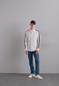 Ung man som bär en vit skjorta med svarta ränder på ärmarna, blå jeans, vita sneakers och en svart keps, står mot en grå vägg.