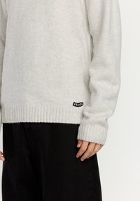 Lys grå genser laget av myk strikket stoff, med ribbestrikket kant og mansjetter, samt et svart logo-merke på nederste venstre side. Kombinert med svarte bukser.
