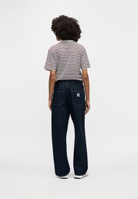 Άτομο με σγουρά μαλλιά, φοράει κοντομάνικο πουκάμισο με ασπρόμαυρες ρίγες, σκούρο τζιν με φαρδιά μπατζάκια και λευκά αθλητικά παπούτσια, στέκεται με την πλάτη προς το μέρος μας.