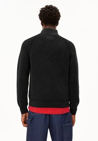 Mann mit kurzen, lockigen Haaren steht mit dem Rücken zur Kamera, trägt einen schwarzen gerippten Pullover über einem roten Hemd und blauen Cargo-Hosen.
