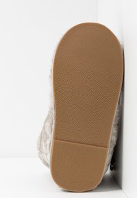 Semelle de chausson en fausse fourrure, avec une semelle extérieure en caoutchouc brun texturé présentant une légère voûte plantaire et un bord supérieur duveteux lisse de couleur gris clair.