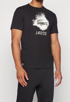 Hombre vistiendo una camisa negra de manga corta con un logo de cocodrilo desgastado en blanco y el texto "LACOSTE" en el pecho.